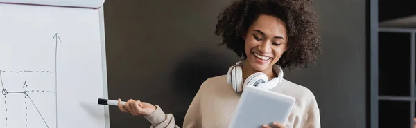 Étudiant afro-américain joyeux avec écouteurs et tablette numérique pointant vers les graphiques sur tableau blanc, bannière — Photo de stock