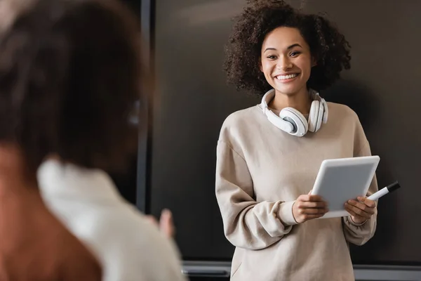 African american student with headphones and digital tablet smiling at classmates on blurred foreground — Stock Photo