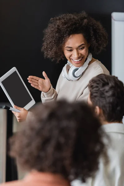 Smiling african american student with headphones showing digital tablet to blurred classmate — Stock Photo