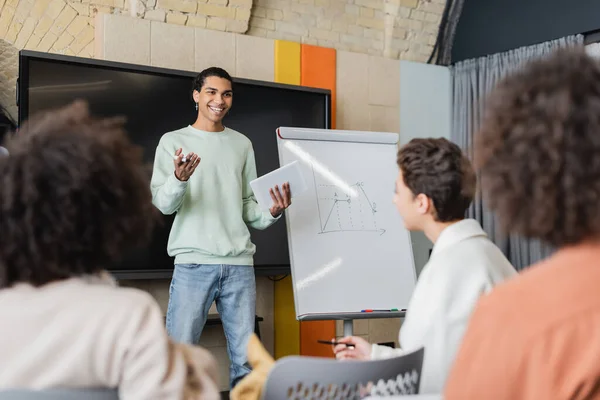 Lächelnder afrikanisch-amerikanischer Student mit digitalem Tablet im Gespräch mit verschwommenen Klassenkameraden neben Whiteboard mit Grafiken — Stockfoto