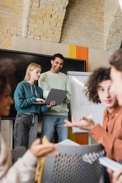 African american student smiling near friend with laptop and blurred interracial classmates talking in classroom — Stock Photo