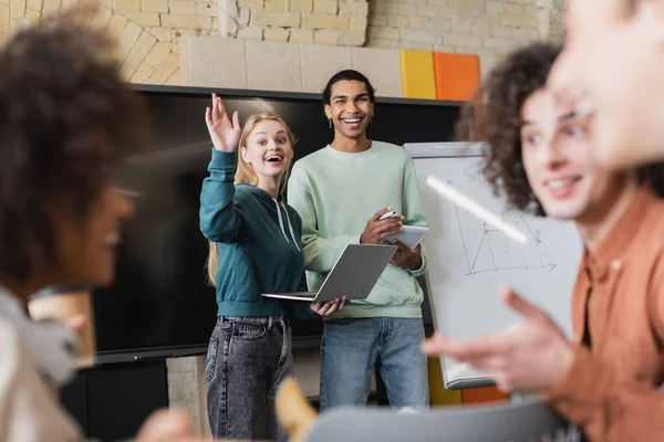 Femme excitée agitant la main près des étudiants multiethniques parlant sur le premier plan flou — Photo de stock