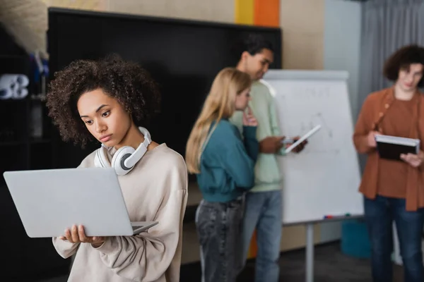 Thoughtful african american woman with headphones looking at laptop near blurred classmates — Stock Photo