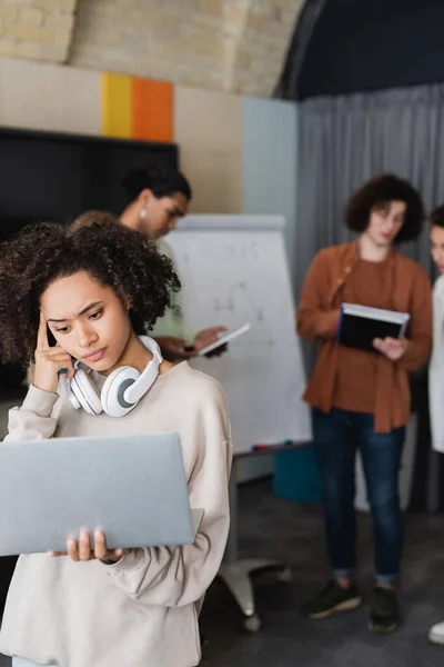 Nachdenkliche afrikanisch-amerikanische Frau mit Laptop und Kopfhörer in der Nähe von verschwommenen interrassischen Studenten — Stockfoto