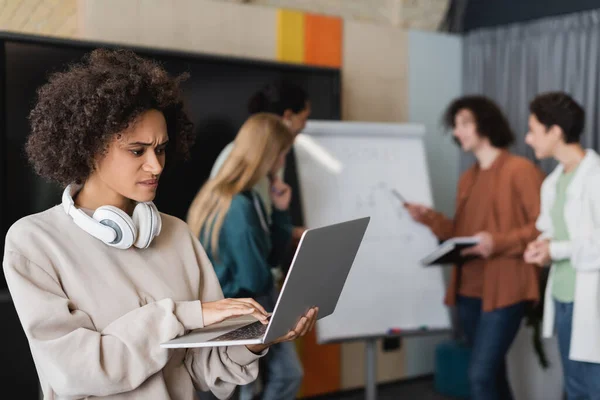Angespannte afrikanisch-amerikanische Frau benutzt Laptop in der Nähe multiethnischer Studenten, die auf verschwommenem Hintergrund sprechen — Stockfoto