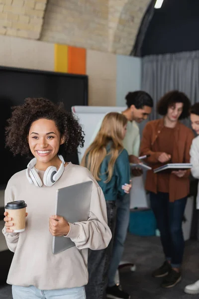 Mujer afroamericana feliz con taza de papel, portátil y auriculares cerca de compañeros de clase en un fondo borroso - foto de stock