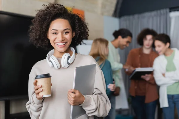 Fröhliche afrikanisch-amerikanische Frau mit Einwegbecher, Kopfhörer und Laptop in der Nähe verschwommener multiethnischer Klassenkameraden — Stockfoto