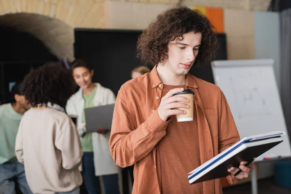 Lockiger Student mit Imbissgetränk, der in der Nähe verschwommener multiethnischer Klassenkameraden auf ein Werbebuch blickt — Stockfoto