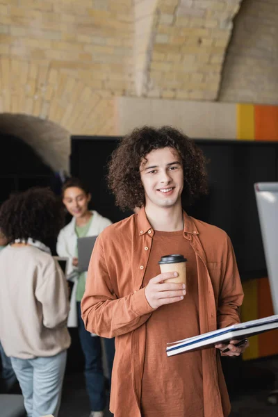 Curly student with takeaway drink and copybooks smiling at camera near women on blurred background — Stock Photo