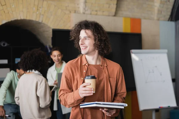 Uomo sorridente con caffè e quaderni che distoglie lo sguardo da amici multietnici offuscati in classe — Foto stock