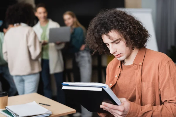 Étudiant bouclé avec copybooks penser près de camarades de classe parler sur fond flou — Photo de stock