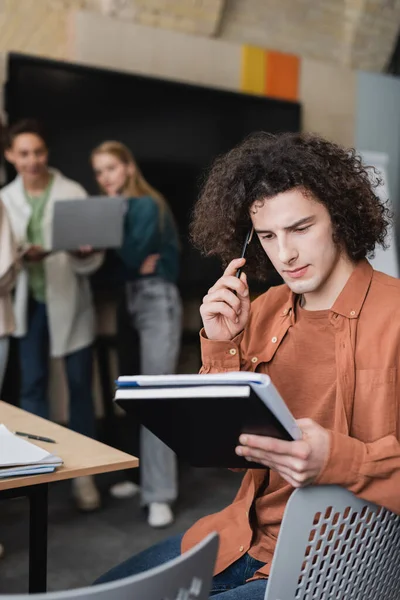 Nachdenklicher Mann hält Stift und Notizbücher in der Hand, während Mitschüler mit Laptop auf verschwommenem Hintergrund reden — Stockfoto