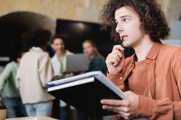 Estudiante reflexivo con copybooks mirando hacia otro lado cerca de compañeros de clase en el auditorio borroso - foto de stock