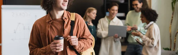 Sorrindo homem com café para ir perto de estudantes inter-raciais olhando para laptop em fundo borrado, banner — Fotografia de Stock