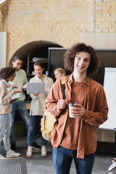 Lockiger Student mit Rucksack und Imbissgetränk in der Nähe verschwommener Freunde, die auf Laptop schauen — Stockfoto