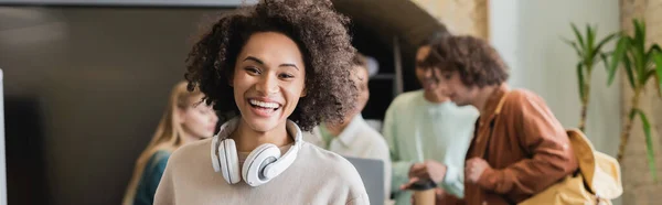 Laughing woman with headphones looking at camera near classmates on blurred background, banner — Stock Photo