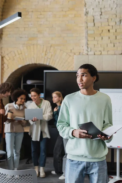 Nachdenkliche afrikanisch-amerikanische Studentin steht mit Notizbuch in der Nähe von Freunden mit Gadgets auf verschwommenem Hintergrund — Stockfoto
