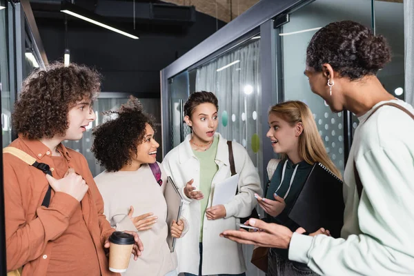 Estudiantes multiculturales asombrados haciendo gestos durante la conversación en el salón de la universidad - foto de stock