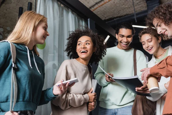 Smiling african american man showing copybook to excited interracial students — Stock Photo