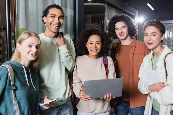 Joyeuse femme afro-américaine avec ordinateur portable regardant la caméra avec ses camarades de classe — Photo de stock