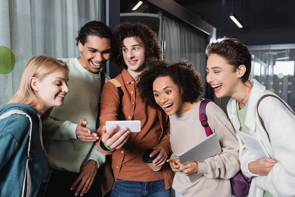 Jeune homme bouclé montrant téléphone portable à des étudiants multiethniques étonnés — Photo de stock