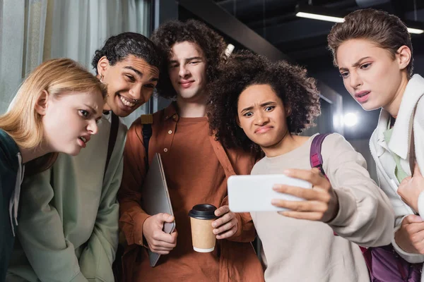 Estudiantes multiétnicos impactados mirando el teléfono móvil en la mano de la mujer afroamericana - foto de stock