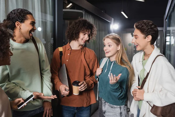 Cheerful multicultural students talking and gesturing in university hallway — Stock Photo