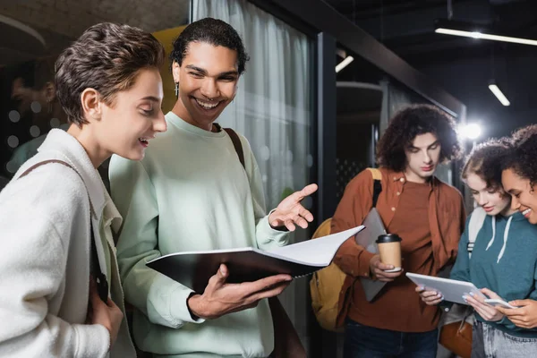 Hombre afroamericano feliz mostrando copybook a mujer cerca de amigos interracial mirando tableta digital - foto de stock