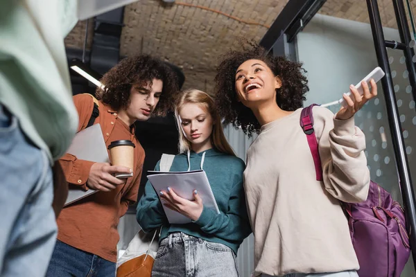 Erstaunt afrikanisch-amerikanische Frau hält Smartphone in der Nähe von Freunden mit Coffee to go und Notizbuch — Stockfoto