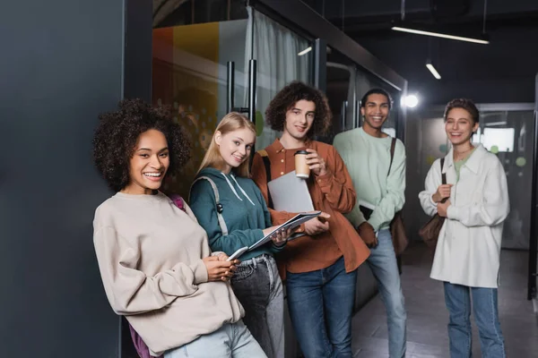 Grupo de positivo interracial estudantes de pé no corredor universitário e sorrindo para a câmera — Fotografia de Stock