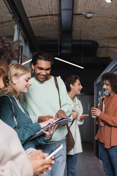 Allegro studente afroamericano che punta il copybook nelle mani del compagno di classe sorridente — Foto stock