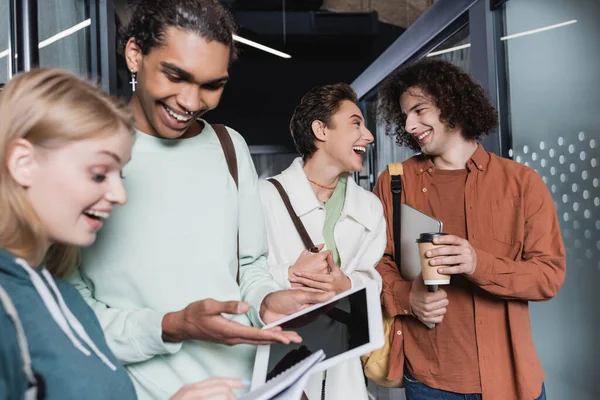 Estudiante americano aricano sonriente apuntando a la tableta digital cerca de compañero de clase sorprendido - foto de stock