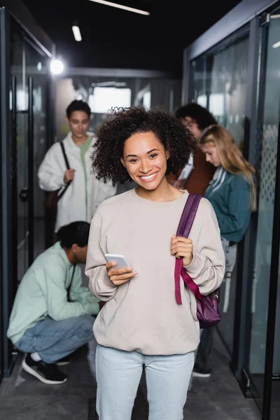Young african american woman with smartphone and backpack smiling at camera near blurred friends — Stock Photo
