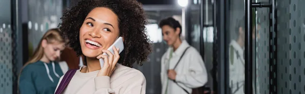 Fröhliche afrikanisch-amerikanische Frau spricht auf Smartphone in der Nähe von Studenten im verschwommenen Flur, Transparent — Stockfoto