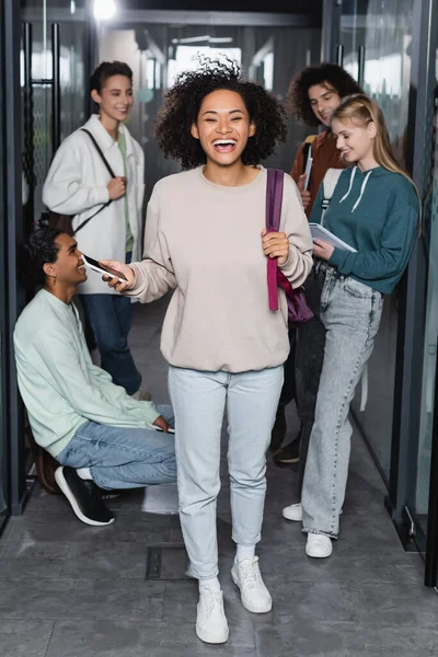 Pleine longueur de rire femme afro-américaine avec smartphone près des amis interraciaux dans le couloir de l'université — Photo de stock