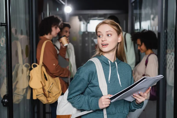 Estudiante sorprendido con mochila y copybook mirando hacia otro lado cerca borrosa compañeros de clase multiétnicos - foto de stock