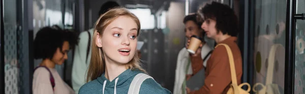 Amazed woman looking away near interracial classmates on blurred background, banner — Stock Photo