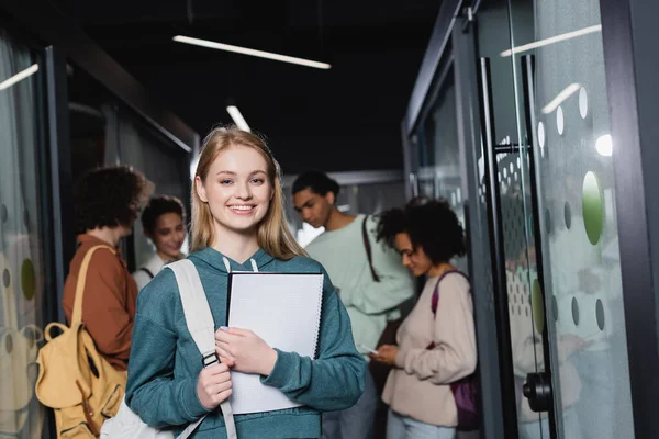 Estudiante alegre con copybook y mochila mirando a la cámara cerca de amigos multiétnicos en fondo borroso - foto de stock