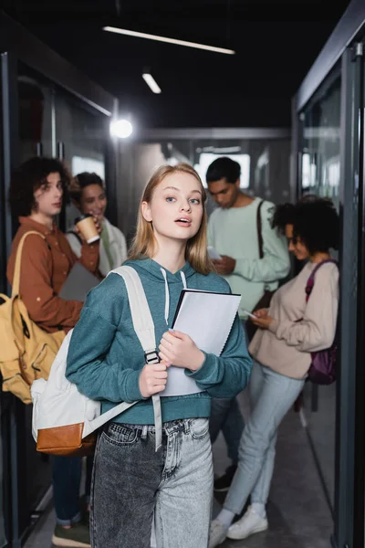 Mujer asombrada con mochila y cuaderno mirando hacia otro lado cerca de estudiantes interracial en pasillo borroso — Stock Photo