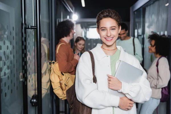 Glückliche junge Frau mit Laptop blickt in die Kamera in der Nähe multiethnischer Freunde auf verschwommenem Hintergrund — Stockfoto