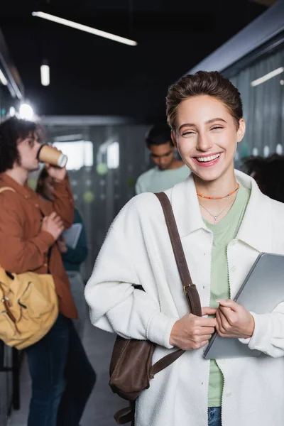 Lachende Frau mit Laptop blickt in die Kamera in der Nähe von interrassischen Studenten im verschwommenen Flur — Stockfoto