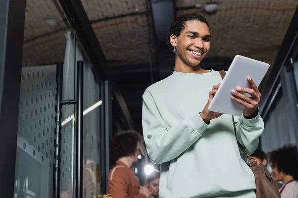 Low angle view of african american student with piercing using digital tablet in hallway of university — Stock Photo