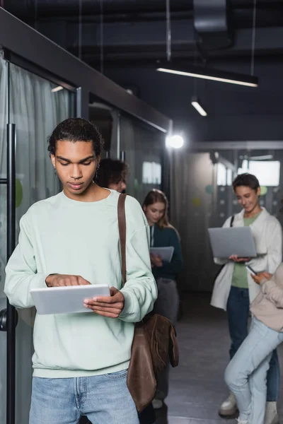 Junger afrikanisch-amerikanischer Mann benutzt digitales Tablet in der Nähe einer verschwommenen Gruppe von Studenten — Stockfoto