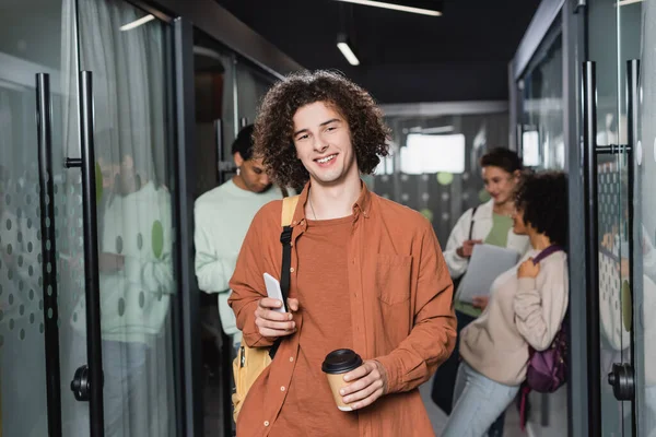 Jovem feliz com café e smartphone olhando para a câmera perto de estudantes multiétnicos no corredor embaçado — Fotografia de Stock