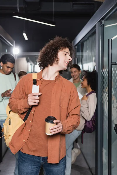Curly student with takeaway drink and backpack looking away near multicultural classmates on blurred background — Stock Photo