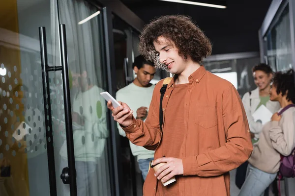 Homme bouclé avec boisson à emporter et téléphone portable près des étudiants multiethniques flous dans le couloir flou — Photo de stock