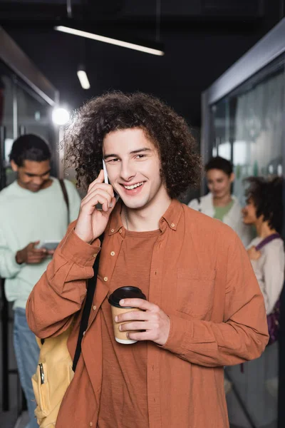Étudiant bouclé et heureux avec tasse en papier parlant sur smartphone près de personnes multiethniques floues — Photo de stock