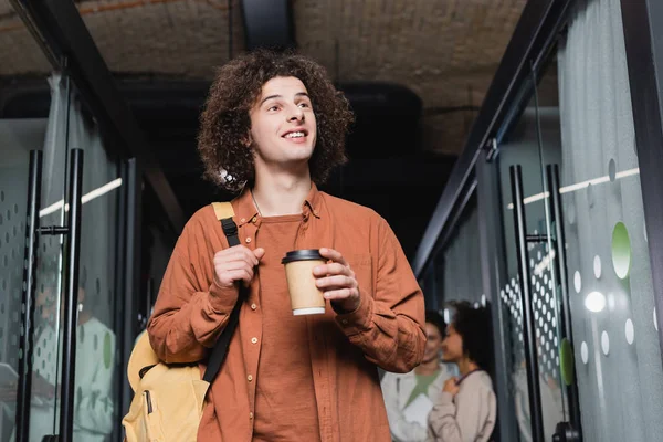 Étudiant bouclé avec café pour aller et sac à dos regardant loin et souriant à l'université — Photo de stock