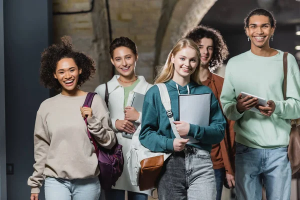 Young and happy interracial students with gadgets smiling at camera in university — Stock Photo