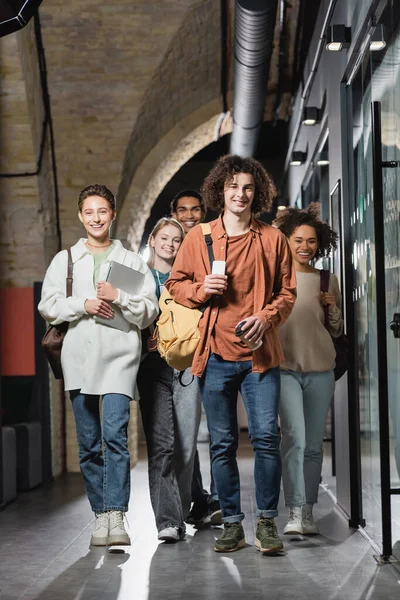 Longitud completa de sonrientes estudiantes interracial con gadgets caminando a lo largo del pasillo en la universidad - foto de stock